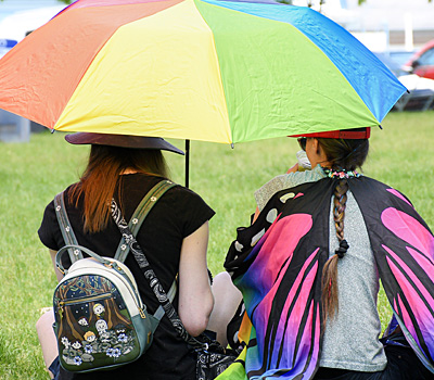 Two people sit on the grass under a rainbow umbrella. One has butterfly wings.