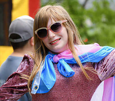 A young person wearing pink sunglasses, a sparkly pink dress, and a blue and pink scarf having fun at an event in the park